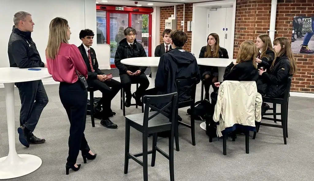 Bright classroom with brick walls, students seated at a round table, and teachers overseeing.