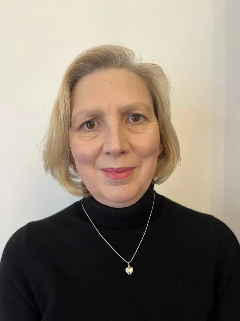 Smiling woman with short blonde hair wearing a black turtleneck and silver heart pendant.